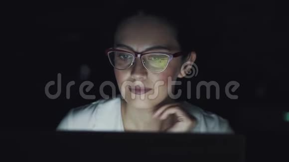 一个年轻的女人晚上在笔记本电脑上工作一个在黑暗的房间里戴眼镜的女商人的肖像视频的预览图