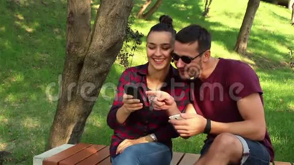 恋爱中的情侣坐在公园的长凳上在智能手机上互相展示照片视频的预览图