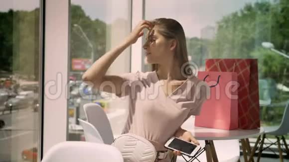 年轻漂亮的女孩白天坐在一家有大窗户的咖啡馆里摸着头发拿着智能手机桌子上有袋子视频的预览图