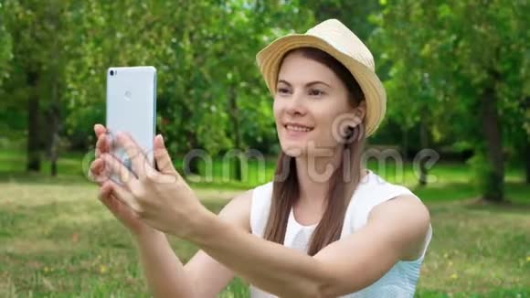 年轻的女大学生穿着白衬衫坐在草地上玩手机自拍视频的预览图