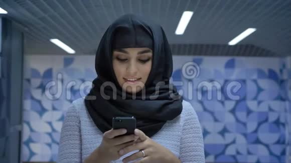 年轻迷人的穆斯林妇女戴着头巾在地下通道的智能手机上输入信息在镜头前观看微笑着宗教视频的预览图
