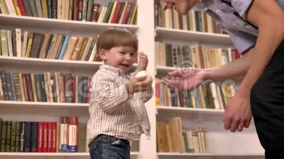 年轻的父亲和他的小儿子玩球微笑快乐书架上有书背景视频的预览图