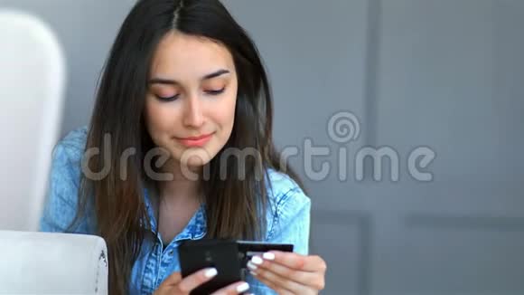 年轻漂亮的女人在家里的客厅里躺在沙发上用信用卡和智能手机上网购物视频的预览图