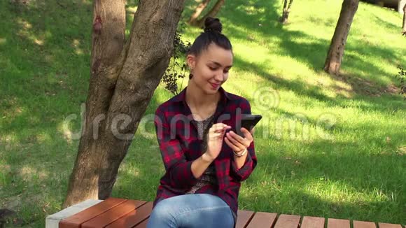 年轻漂亮的女孩坐在公园的长凳上在手机里浏览社交网络磁带视频的预览图