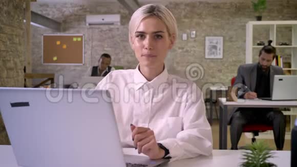 年轻的快乐女商人坐在办公室的笔记本电脑前跳舞同事们正在与技术工作建立联系视频的预览图