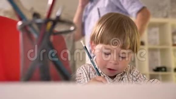 小男孩在纸上画画微笑着当他的父亲在背景上打电话时他高兴地坐在现代的椅子上视频的预览图