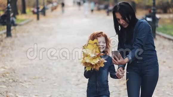 一位美丽的妈妈和她可爱的小女儿在秋天的公园里散步他们在智能手机上自拍笑着视频的预览图