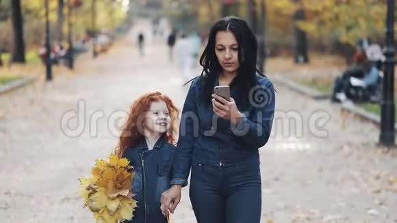 一位美丽的妈妈和她可爱的小女儿在秋天的公园里散步他们在智能手机上自拍笑着视频的预览图