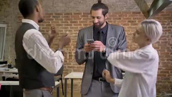三个不同种族的工人在办公室跳舞看智能手机公司聚会的概念视频的预览图
