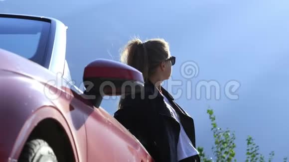 一个穿着皮夹克和太阳镜的年轻时髦黑发女孩站在一辆红色敞篷车旁边视频的预览图