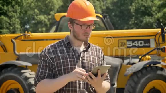 年轻建筑工人戴着头盔拿着平板电脑皱着眉头向前看深思熟虑背景上的拖拉机的肖像视频的预览图