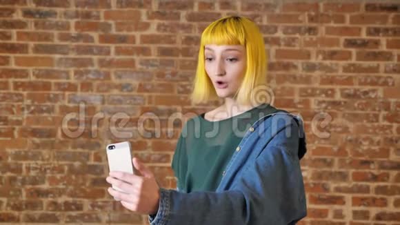 黄色头发的年轻女子通过视频聊天与某人交谈站在砖背景附近视频的预览图