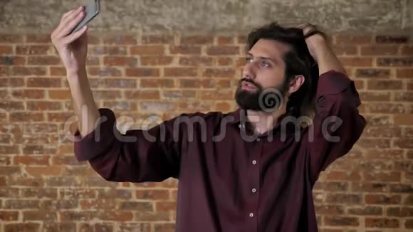 年轻有魅力的黑发男子带着胡须正在智能手机上自拍触摸头发砖背景交流视频的预览图