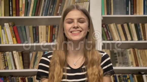 年轻可爱的金发女郎正在镜头前看着微笑着举手背景上的图书馆视频的预览图