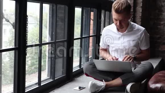 一个年轻人坐在窗台上用笔记本电脑和智能手机工作视频的预览图
