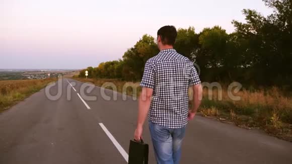 穿格子衬衫的人手里拿着一个黑色公文包沿着沥青路走着视频的预览图