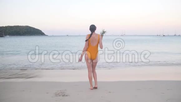 穿着黄色泳衣的女人在日落的沙滩上和菠萝跳舞视频的预览图