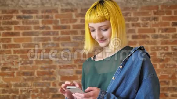 年轻不寻常的女人一头黄发拿着电话看着摄像机微笑着站在砖墙附近视频的预览图