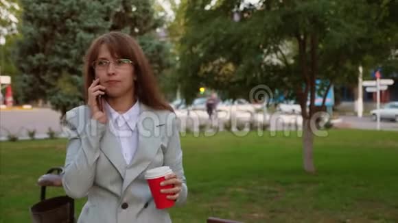 女商人戴着浅色西装眼镜在公园里散步喝咖啡在智能手机上聊天视频的预览图