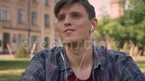 年轻的帅哥坐在公园的草地上用耳机听音乐唱歌放松概念继续前进视频的预览图