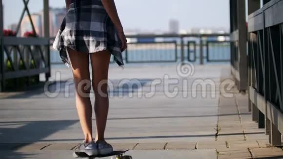 在一个阳光明媚的夏天一个美丽苗条的女孩沿着河堤骑着滑板视频的预览图