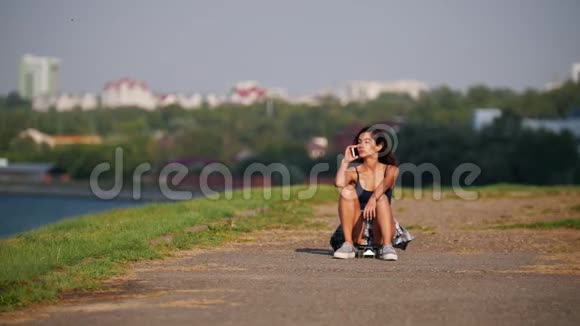 美丽的女孩坐在滑板上在岸上打电话视频的预览图