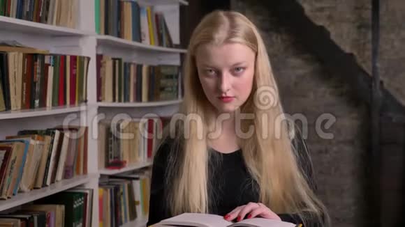 年轻漂亮的金发女孩涂着粉红色口红正在看书看着相机微笑着在后台看图书馆视频的预览图