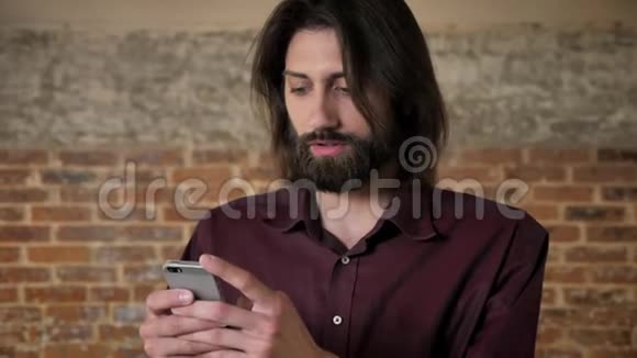 年轻迷人的黑发男子蓄着胡须在智能手机上观看在镜头下观看砖背景交流视频的预览图