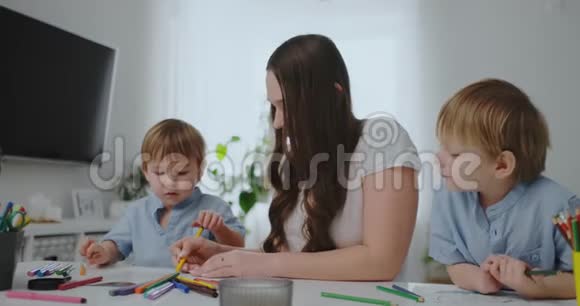 一位年轻的母亲和两个孩子坐在一张白色的桌子旁在纸上画着彩色铅笔帮助做家庭作业视频的预览图