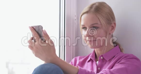 漂亮的金发女孩坐在窗户上手里拿着智能手机视频的预览图