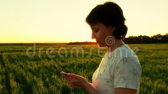 一个年轻有魅力的女人正拿着一部智能手机在夕阳的背景下在一片绿色的草坪上慢动作视频的预览图