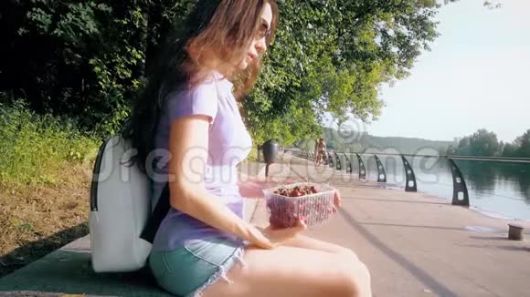 美丽的黑发少女在城市公园堤岸吃甜樱桃视频的预览图