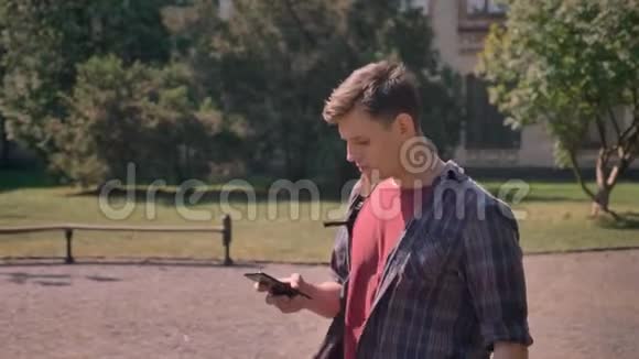 年轻的帅哥正在夏天的白天穿过公园敲着智能手机交流理念视频的预览图