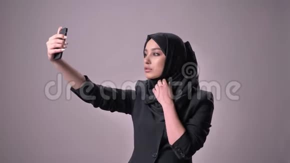 年轻漂亮的穆斯林女孩戴着头巾在智能手机上自拍看照片交流观念灰色视频的预览图