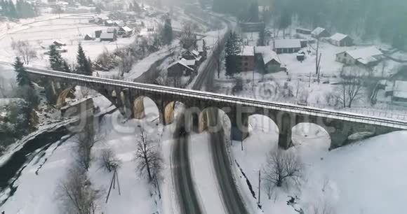 一辆汽车在一个白雪覆盖的村庄4K超高清的铁路桥下通过的鸟瞰图视频的预览图