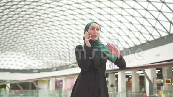 戴着头巾和背包的漂亮穆斯林女人站在一家购物中心打电话视频的预览图