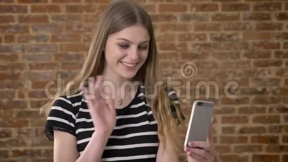 年轻快乐的金发女郎在智能手机上视频聊天挥手问好交流理念砖背景视频的预览图