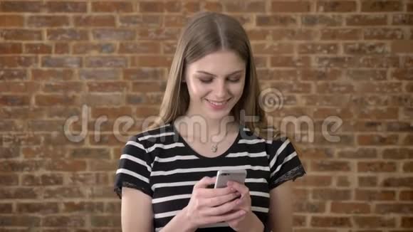 年轻可爱的金发女郎正在智能手机上输入信息看着相机微笑着交流理念砖背景视频的预览图