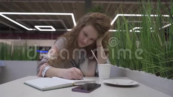 年轻漂亮的红发女人坐在咖啡馆里写在笔记本上视频的预览图