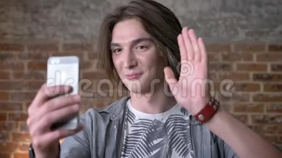 年轻的长发男子带着鼻环在智能手机上制作视频挥手问好展示和平的姿态交流视频的预览图