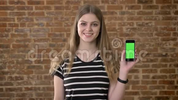 年轻快乐的金发女郎正在展示智能手机的绿色屏幕指向它沟通概念砖背景视频的预览图