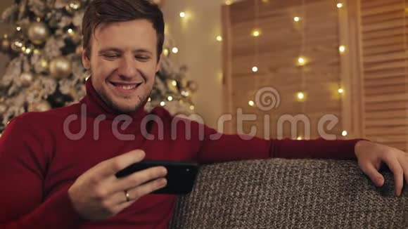 迷人的男人使用智能手机坐在沙发上发信息微笑在装饰好的公寓附近他在看录像视频的预览图