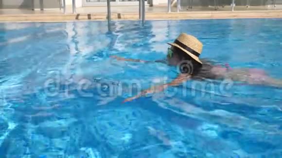 年轻漂亮的女孩戴着太阳镜和帽子在游泳池游泳在阳光明媚的日子里女人在清澈的温水中放松夏季视频的预览图
