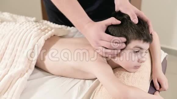 一位男性理疗师按摩师为躺在按摩床上的一个微笑的小男孩做一个疗愈放松的按摩头视频的预览图