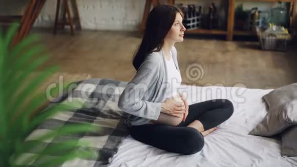 可爱的期待妈妈闭上眼睛抚摸她的肚子感觉婴儿坐在现代房子的床上视频的预览图