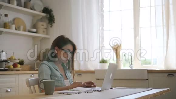 年轻的女性自由职业者ig眼镜工作在笔记本电脑和笔记在明亮的厨房视频的预览图