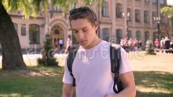 年轻的帅哥把黑色皮夹克放在一个肩膀上拿着背包时髦的学生站在公园附近视频的预览图