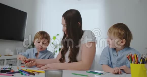 一位年轻的母亲和两个孩子坐在一张白色的桌子旁在纸上画着彩色铅笔帮助做家庭作业视频的预览图