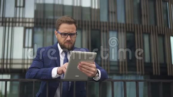 戴着眼镜和蓝色西装的商人在现代办公楼附近发布新平板电脑的新闻视频的预览图
