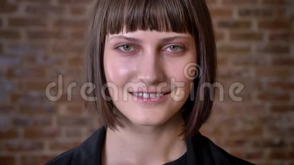 年轻的女人剪短头发绿眼睛看着相机微笑着在砖的背景上与世隔绝视频的预览图